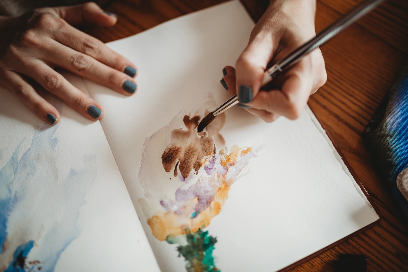 Hands painting soft watercolor shapes in a sketchbook on a wooden table